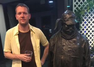 Justin Kern of the Museum of the Unintentional stands, with a drink in hand, next to a bronze statue of a person in a hunting cap with ear flaps.